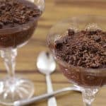 Two elegant glass dessert cups filled with chocolate mousse, topped with chocolate shavings, are presented on a wooden surface. Two silver spoons are placed next to the cups, ready for use.