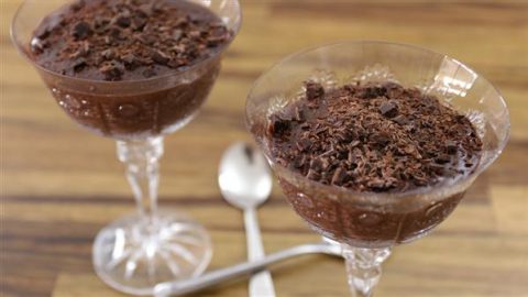 Two elegant glass dessert cups filled with chocolate mousse, topped with chocolate shavings, are presented on a wooden surface. Two silver spoons are placed next to the cups, ready for use.