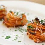 Three stacks of thinly sliced sweet potatoes, garnished with grated cheese and finely chopped herbs, are arranged on a white plate. The sweet potatoes appear roasted and are layered neatly. The dish is presented on a wooden table.