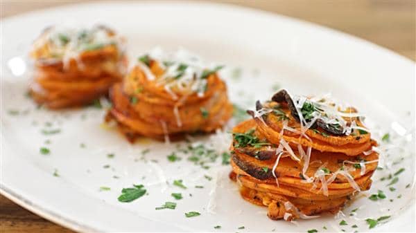 Three stacks of thinly sliced sweet potatoes, garnished with grated cheese and finely chopped herbs, are arranged on a white plate. The sweet potatoes appear roasted and are layered neatly. The dish is presented on a wooden table.