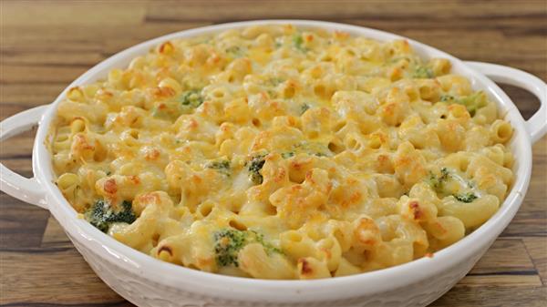 A round white casserole dish filled with baked macaroni and cheese, mixed with small pieces of broccoli, showing a golden browned cheese crust on top. The dish is placed on a wooden surface.