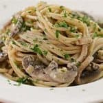 A white plate filled with creamy pasta topped with sliced mushrooms and garnished with chopped parsley. The pasta is mixed with a rich, smooth sauce, and is served on a wooden table.