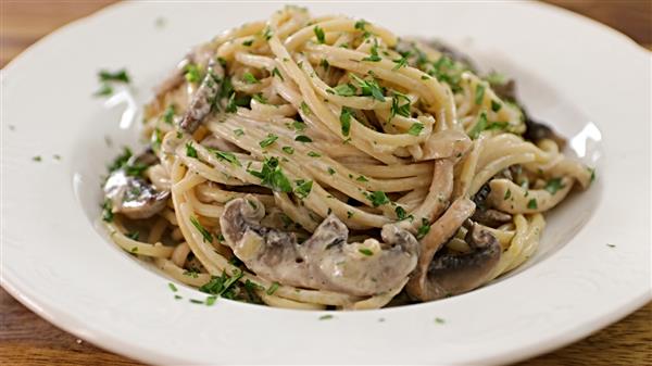 A white plate filled with creamy pasta topped with sliced mushrooms and garnished with chopped parsley. The pasta is mixed with a rich, smooth sauce, and is served on a wooden table.