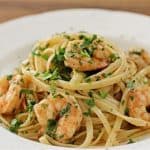 A white plate of linguine pasta topped with cooked shrimp, finely chopped parsley, and herbs. The dish is garnished with a light sauce and fresh herbs, creating an appetizing presentation on a wooden table background.