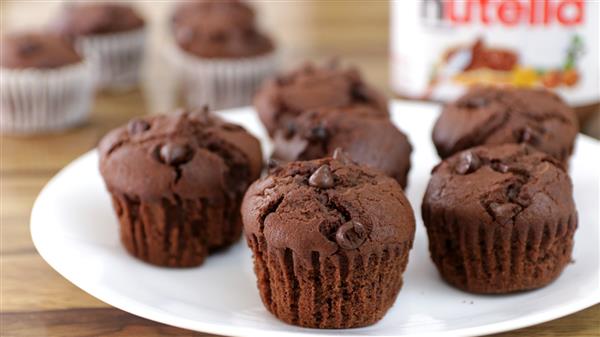 A white plate filled with chocolate muffins, with visible chocolate chips, is placed on a wood surface. In the background, there are three more muffins and a jar of Nutella, slightly blurred. The muffins appear freshly baked and moist.