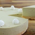 A whole matcha cheesecake with a slice being served. The cheesecake has a light green matcha filling on a biscuit crust, and is decorated with eight evenly spaced dollops of white whipped cream on top. It rests on a wooden surface.