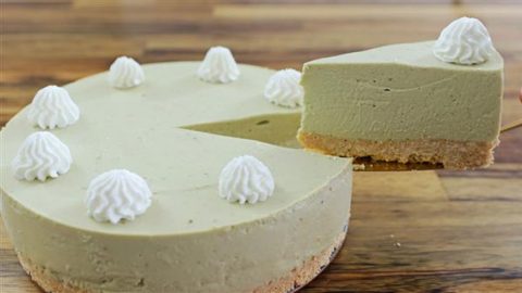 A whole matcha cheesecake with a slice being served. The cheesecake has a light green matcha filling on a biscuit crust, and is decorated with eight evenly spaced dollops of white whipped cream on top. It rests on a wooden surface.