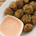 A white plate filled with round, golden-brown falafel balls with a side of pink dipping sauce in a small, white bowl. The plate is placed on a wooden surface.