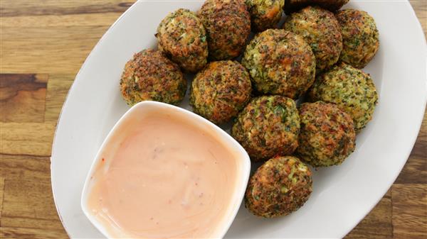 A white plate filled with round, golden-brown falafel balls with a side of pink dipping sauce in a small, white bowl. The plate is placed on a wooden surface.