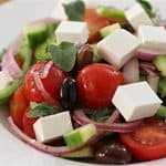 A close-up of a fresh Greek salad featuring cherry tomatoes, cucumber slices, red onion rings, black olives, and cubes of feta cheese, garnished with fresh herbs, served on a white plate.