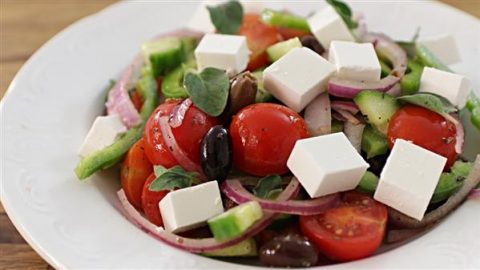 A close-up of a fresh Greek salad featuring cherry tomatoes, cucumber slices, red onion rings, black olives, and cubes of feta cheese, garnished with fresh herbs, served on a white plate.