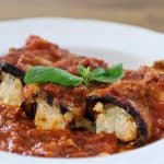 A white plate holds two eggplant rollatini, stuffed with ricotta cheese and topped with marinara sauce. A single basil leaf garnishes the dish, adding a touch of green to the rich red sauce. The rollatini are nestled in a generous pool of marinara sauce.
