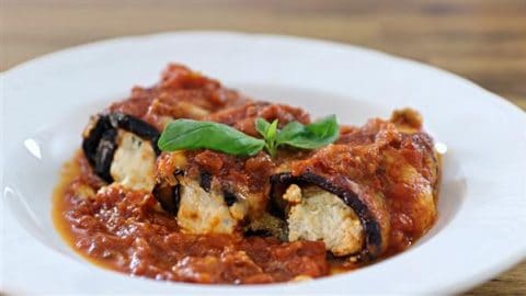 A white plate holds two eggplant rollatini, stuffed with ricotta cheese and topped with marinara sauce. A single basil leaf garnishes the dish, adding a touch of green to the rich red sauce. The rollatini are nestled in a generous pool of marinara sauce.