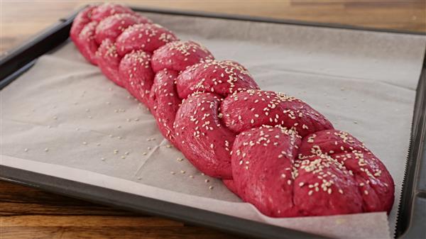 A red braided loaf of bread sprinkled with sesame seeds rests on a baking sheet lined with parchment paper, placed on a wooden surface.