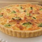 A freshly baked quiche with a golden brown crust sits on a white plate. The filling includes broccoli and appears cheesy, with browned spots on top. The background is a wooden table.