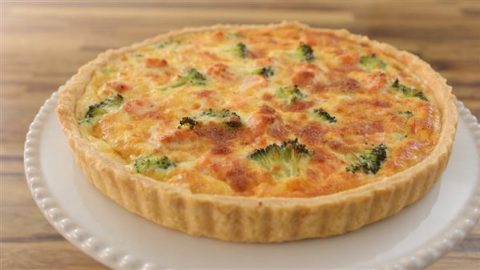 A freshly baked quiche with a golden brown crust sits on a white plate. The filling includes broccoli and appears cheesy, with browned spots on top. The background is a wooden table.