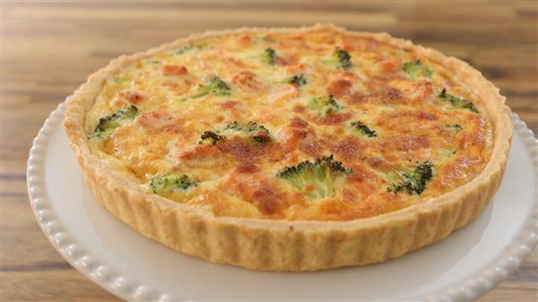 A freshly baked quiche with a golden brown crust sits on a white plate. The filling includes broccoli and appears cheesy, with browned spots on top. The background is a wooden table.