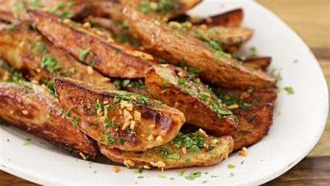 A plate of roasted potato wedges garnished with chopped herbs and minced garlic. The wedges are crispy and golden brown, presented on a white dish.