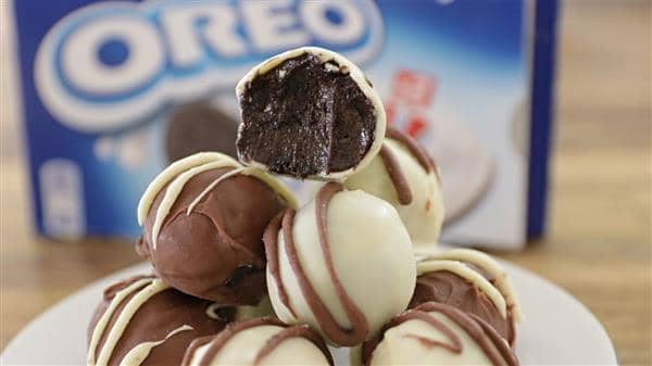 A plate of Oreo truffles is stacked in a pyramid, with a biten one showcased at the top. The truffles are coated in white and milk chocolate drizzles, with an Oreo package in the blurred background.