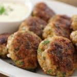 A plate of browned, flavorful meatballs garnished with fresh herbs, served alongside a small bowl of creamy white dipping sauce. The meatballs are arranged on a white oval plate, emphasizing their golden-brown exterior.