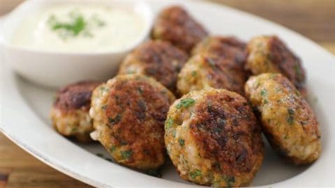 A plate of browned, flavorful meatballs garnished with fresh herbs, served alongside a small bowl of creamy white dipping sauce. The meatballs are arranged on a white oval plate, emphasizing their golden-brown exterior.