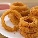 A white plate filled with golden-brown, crunchy onion rings is placed on a wooden table. A small white dish containing red dipping sauce is situated in the background. The onion rings are stacked in multiple piles.
