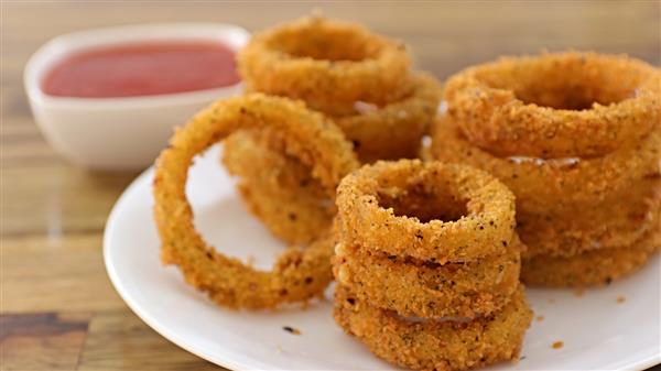 A white plate filled with golden-brown, crunchy onion rings is placed on a wooden table. A small white dish containing red dipping sauce is situated in the background. The onion rings are stacked in multiple piles.