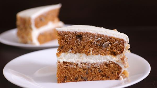 A close-up of a slice of carrot cake on a white plate. The cake has two layers of moist, brown cake with visible nuts and is filled and topped with white cream cheese frosting. Another slice of cake is blurred in the background.