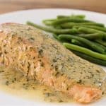 A white plate featuring a cooked salmon fillet topped with a creamy herb sauce, accompanied by a serving of steamed green beans. The dish is presented on a wooden surface.