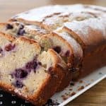 A rectangular loaf of blueberry bread is partially sliced on a white platter. The bread has a golden brown crust and is dusted with powdered sugar. The interior is light and fluffy, studded with juicy blueberries. The platter rests on a wooden surface.