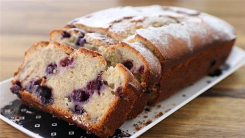 A rectangular loaf of blueberry bread is partially sliced on a white platter. The bread has a golden brown crust and is dusted with powdered sugar. The interior is light and fluffy, studded with juicy blueberries. The platter rests on a wooden surface.