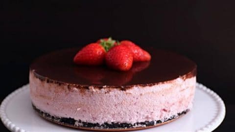 A round strawberry mousse cake with a chocolate ganache top layer, garnished with three fresh strawberries, sits on a white cake stand against a dark background. The cake has a visible layer of crushed cookie crust at the bottom.