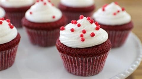 Red velvet cupcakes topped with swirls of white frosting and red decorative sprinkles are arranged on a white serving plate.