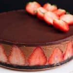 A round chocolate mousse cake with a dark chocolate glaze on top, decorated with halved strawberries arranged in a fan shape. Halved strawberries are also embedded around the side. The cake sits on a white plate against a dark background.