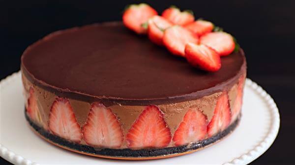 A round chocolate mousse cake with a dark chocolate glaze on top, decorated with halved strawberries arranged in a fan shape. Halved strawberries are also embedded around the side. The cake sits on a white plate against a dark background.