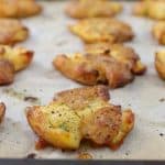 A baking sheet lined with parchment paper holds several pieces of crispy, golden-brown smashed potatoes seasoned with herbs and pepper. The potatoes are slightly flattened with a rugged texture and appear freshly baked.