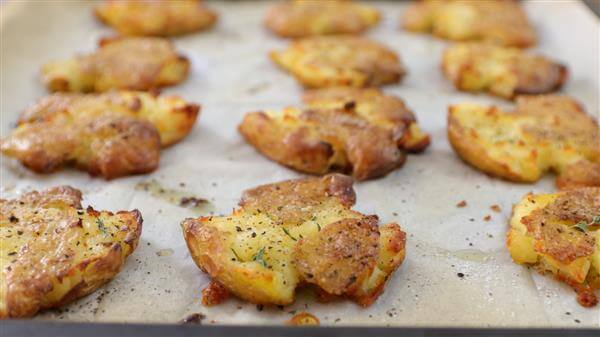 A baking sheet lined with parchment paper holds several pieces of crispy, golden-brown smashed potatoes seasoned with herbs and pepper. The potatoes are slightly flattened with a rugged texture and appear freshly baked.