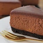 A slice of rich chocolate cheesecake with a dark chocolate crust sits on a white plate. A golden fork is placed next to the cheesecake slice. The background features another whole cheesecake out of focus.