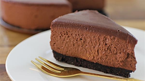 A slice of rich chocolate cheesecake with a dark chocolate crust sits on a white plate. A golden fork is placed next to the cheesecake slice. The background features another whole cheesecake out of focus.