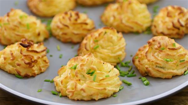 A plate of golden-brown duchess potatoes garnished with finely chopped chives. The potatoes are piped into neat, swirled mounds, showcasing a crispy exterior with a fluffy interior. They are artfully arranged on the plate, ready to be served.