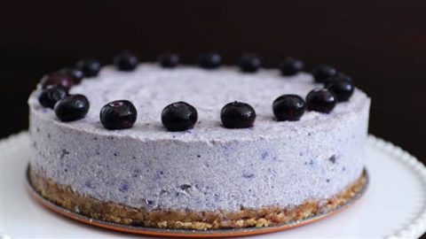 A round blueberry cheesecake with a crumbly crust sits on a white plate. The cheesecake is topped with a ring of fresh blueberries, placed evenly around the edge. The dessert has a smooth, creamy texture with a light purple color. The background is dark.