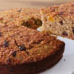 A slice of dense, golden-brown oat cake with visible pieces of dried fruit being lifted on a cake server. The rest of the cake sits on a white pedestal plate on a wooden surface.