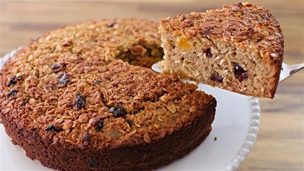 A slice of dense, golden-brown oat cake with visible pieces of dried fruit being lifted on a cake server. The rest of the cake sits on a white pedestal plate on a wooden surface.