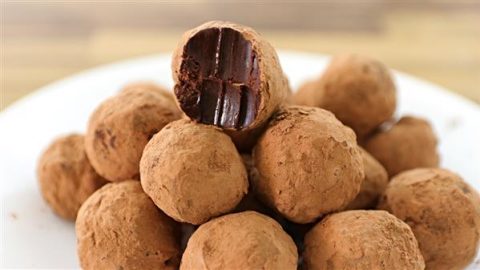 A plate is piled with round chocolate truffles coated in cocoa powder. The truffle at the top of the pile has a bite taken out, revealing a rich, creamy chocolate interior. The background is slightly blurred.