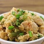 A bowl of cauliflower rice mixed with pieces of chicken and green beans, garnished with chopped green onions, is placed on a wooden surface. The dish has a textured, crumbly appearance with visible chunks of chicken and vegetables.