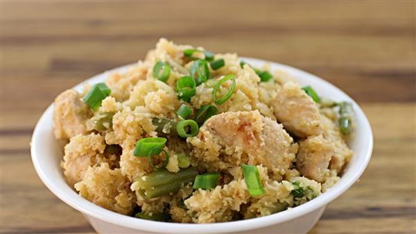 A bowl of cauliflower rice mixed with pieces of chicken and green beans, garnished with chopped green onions, is placed on a wooden surface. The dish has a textured, crumbly appearance with visible chunks of chicken and vegetables.