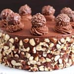 A chocolate cake adorned with chopped hazelnuts on its sides sits on a white cake stand. The cake is topped with swirls of chocolate frosting, each holding a Ferrero Rocher chocolate, adding an elegant touch to the decadent dessert.