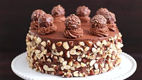 A chocolate cake adorned with chopped hazelnuts on its sides sits on a white cake stand. The cake is topped with swirls of chocolate frosting, each holding a Ferrero Rocher chocolate, adding an elegant touch to the decadent dessert.