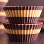 Close-up of stacked chocolate peanut butter cups with alternating layers of chocolate and peanut butter. The cups have a ribbed texture and are set against a blurred, dark background, with additional cups visible in the background.