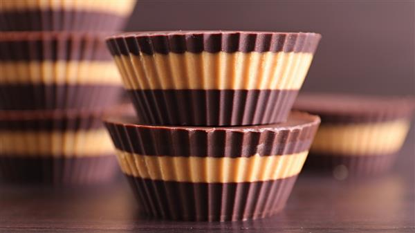 Close-up of stacked chocolate peanut butter cups with alternating layers of chocolate and peanut butter. The cups have a ribbed texture and are set against a blurred, dark background, with additional cups visible in the background.